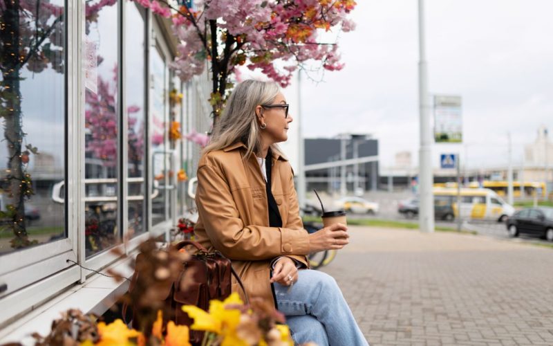 a stylish pensioner sits on a bench next to a store with a mobile phone in her hands and looks into the distance.
